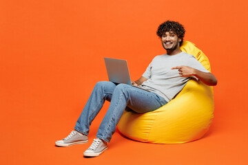 Full body young happy IT Indian man wears t-shirt casual clothes sit in bag chair hold use work...
