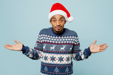 Young man wears knitted sweater Santa hat posing spread hands shrugging shoulders looking puzzled isolated on plain pastel blue background. Happy New Year 2024 celebration Christmas holiday concept.