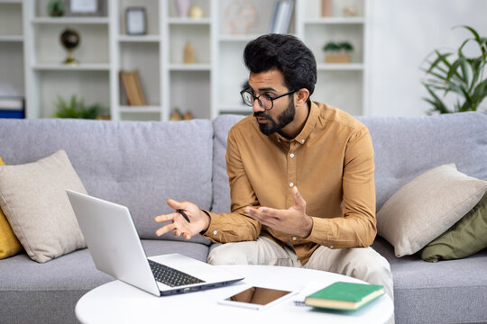 Indian young man businessman, freelancer holding a meeting online. Sitting on the couch and communicating via video call on a laptop.
