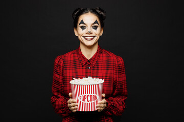 Young fun woman with Halloween makeup face art mask wear clown costume red dress hold in hand pop corn watch movie film isolated on plain black background studio portrait. Scary holiday party concept.