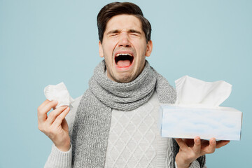 Young sad ill sick man wear gray sweater scarf sneezing hold box of paper napkins cry sneeze isolated on plain blue background studio. Healthy lifestyle disease treatment cold season recovery concept. - obrazy, fototapety, plakaty