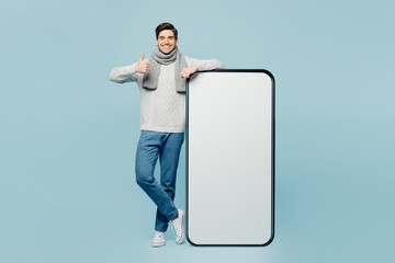 Full body young ill sick man wear gray sweater scarf big blank screen mobile cell phone show thumb up isolated on plain blue background. Healthy lifestyle disease virus cold season recovery concept.