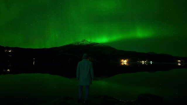 Man standing on a rock beneath a green aurora in October, gazing across a lake and the mountainous landscape.