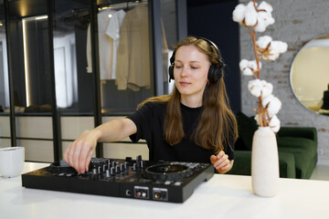 A young woman mixes tracks on a DJ controller for a set while sitting in a room. The concept of...