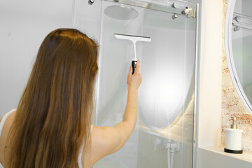 A young woman cleans the glass walls of the shower with a scraper from water drops and plaque in...