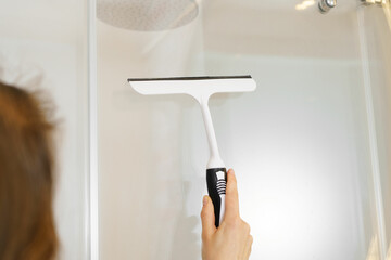 Close-up of a young woman using a scraper to remove drops from a glass shower and lime scale. The concept of cleaning in the apartment and in the bathroom - obrazy, fototapety, plakaty