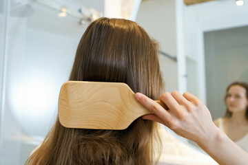 Fototapeta premium Close-up of a young brown-haired woman combing her hair with a wooden comb in the bathroom, rear view. The concept of hair cosmetics and products for shine