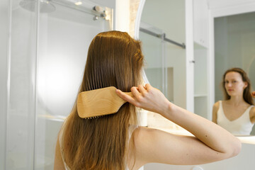 Fototapeta premium Young woman combs her shiny hair with a wooden comb, rear view. Concept of daily body care routine, hair cosmetics