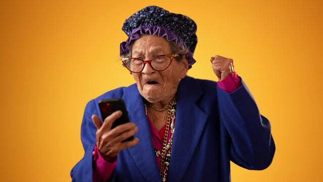 Funny toothless elderly senior old woman with wrinkled skin looking at phone having great happy success winner gesture isolated on yellow background. People emotions, lifestyle concept.