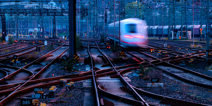 Railway panorama with leaving train in early morning twilight at blue hour. Tracks, oververhead lines, switches at Hagen main station in Ruhr basin, Germany. Transportation technology for passengers.