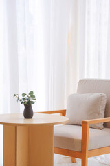 Gray cushion on armchair with ceramic vase on round wooden desk near white curtain inside of modern coffee shop in beige tone style and vertical frame
