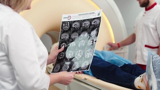In a modern clinic, a female radiologist explains the good results of a CT scan of a young patient, observes and analyzes the CT scan next to a modern CT scanner.