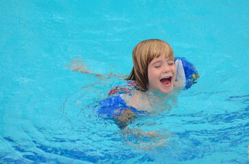 Fototapeta na wymiar A little boy learns to swim in the pool. Child in swimming pool floating on inflatable mattress. Kids swim. Kid l having fun on family summer vacation in resort