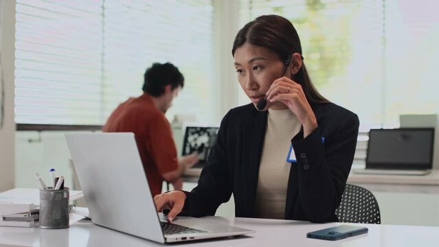 Medium shot of busy female call centre specialist communicating with client via headset while sitting at laptop in office