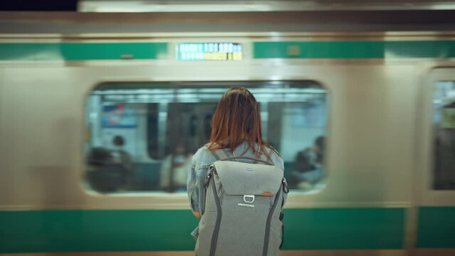 Rear view of young woman with backpack while waiting at platform of subway train station looking at passing tram, slow motion, waiting for a trip.