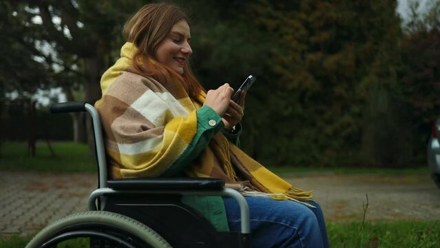 Young woman in bright stylish clothes with physical disability uses a mobile phone, reads news, communicates on social networks sitting in a wheelchair in park in autumn nature. High quality photo
