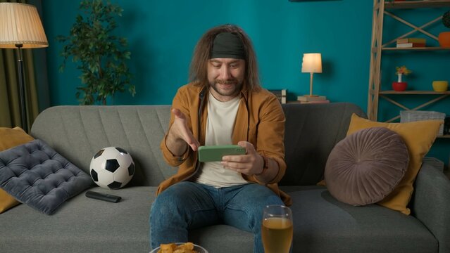In the picture a middle aged man is sitting on a sofa in the living room. He is holding a phone and looking gesticulating on it something. Next to him lies a ball and beer with chips. He is attentive