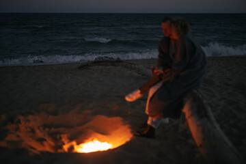 Couple sitting together near campfire on the beach, focus on the sea