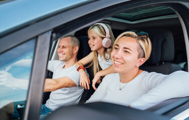 Cheerful young traditional family has a auto journey in modern car. Mother, father and daugher with headphones. Safety riding car and traveling concept image.