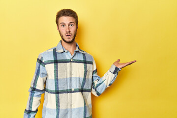 Young Caucasian man on a yellow studio background impressed holding copy space on palm.