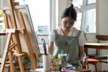 Happy young Asian painter woman Mix oil paints on a paint tray and prepare them for painting on canvas in the painting studio.