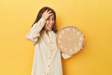 Woman holding clock on yellow backdrop being shocked, she has remembered important meeting.