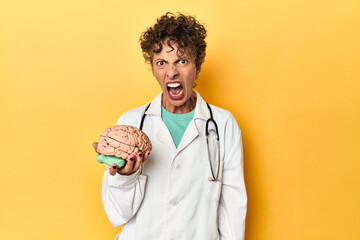 Doctor holding a brain model on yellow studio screaming very angry and aggressive.