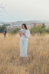Young woman dressed in white standing reading a book in a meadow at sunset