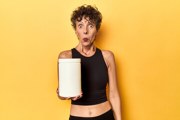 MIddle aged athlete woman holding protein supplement on yellow shrugs shoulders and open eyes...