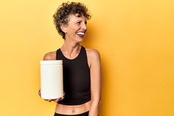 MIddle aged athlete woman holding protein supplement on yellow looks aside smiling, cheerful and...