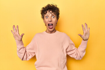 Mid-aged caucasian woman on vibrant yellow receiving a pleasant surprise, excited and raising hands.