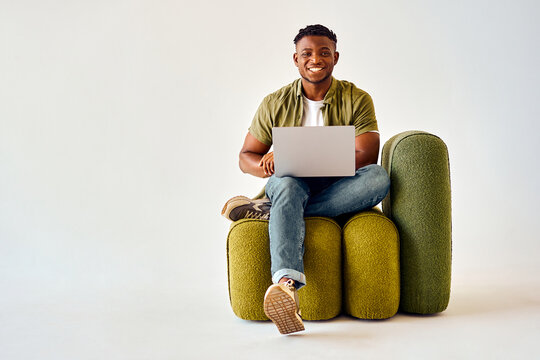 Happy african american guy sitting on design chair and holding wireless laptop. Male freelancer in casual attire looking at camera with toothy smile over white background.