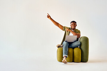 Recommendation of quality device. Full size young afro man pointing with index fingers on empty space of white studio background. Male freelancer holding modern laptop on knees and smiling at camera.
