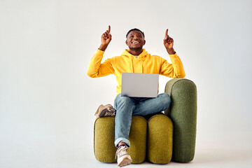Cheerful african american man in yellow hoodie and jeans sitting on green design chair with...