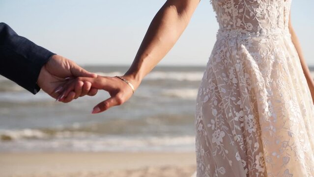 Groom gently holding bride's hand, newlyweds walking at the sandy beach . Pretty woman in lace dress and man in suit going at the seaside. Just married couple enjoy wedding day. Close-up.