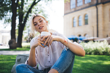Portrait of beautiful millennial woman with takeaway cup looking at camera while listening audio book during leisure pastime in summer park, charming blondie hipster girl in earphones posing