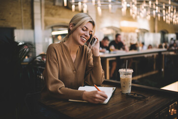 Cheerful female student making informative notes while calling via cellphone device for discussing education information, joyful blonde woman with textbook making positive phone conversation in cafe