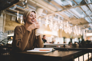Happy Caucasian woman with notebook enjoying positive cellular communication via mobile technology during coffee time in cafe interior, cheerful millennial user calling via smartphone device
