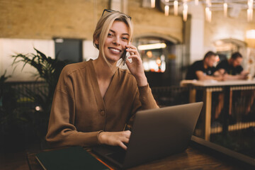 Portrait of cheerful female copywriter using cellphone for call and talk about laptop technology coworking space, happy woman phoning via smartphone device for discussing web communication on netbook