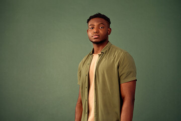 Confidence and pride. Front view of african young man with serious facial expression standing over green studio background. Handsome male in casual khaki shirt controlling emotions and mimics. - obrazy, fototapety, plakaty