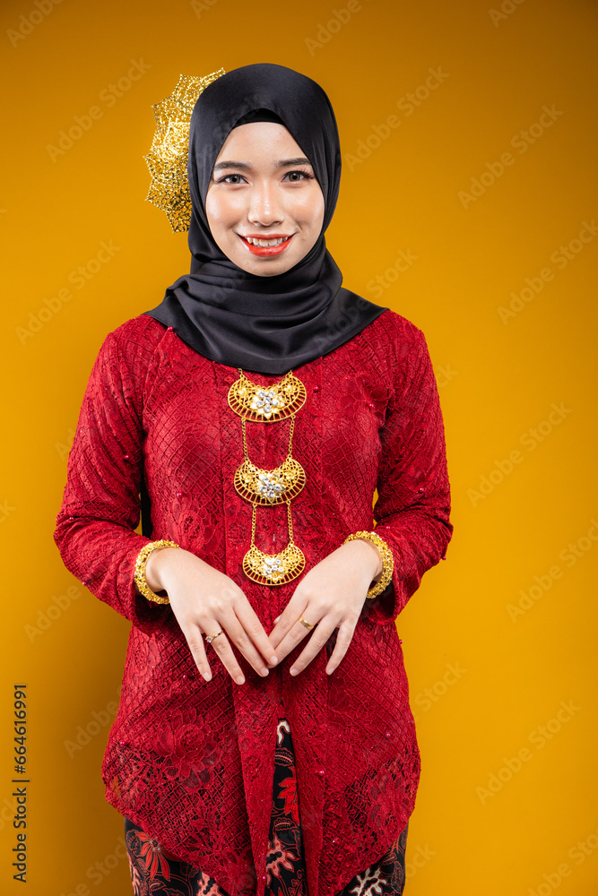 Wall mural Muslim woman wearing traditional wear red kebaya and hijab isolated on yellow background. Idul Fitri and hijab fashion concept.