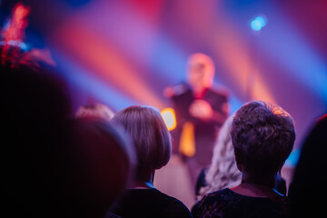 a scene from a public event. The focus is on the back of the heads of an audience, blurred in the...