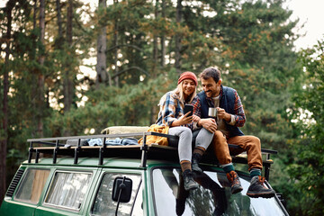 Happy couple using mobile phone while sitting on roof of their camper trailer in nature.