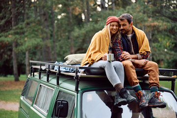 Carefree couple wrapped in blanket relaxing on top of their camping van in nature.