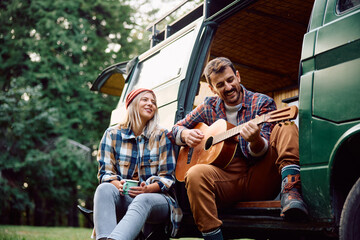 Happy man playing acoustic guitar during camping trip with his girlfriend in woods.