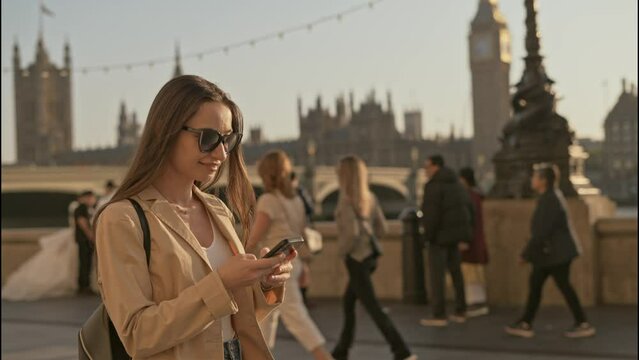 Young tourist woman using smartphone while walking along  Thames River with Big Ben Palace of Westminster background view, on a beautiful sunny morning