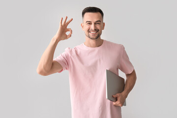 Happy young man with laptop showing OK on light background