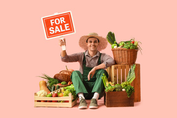 Male farmer with fresh vegetables and FOR SALE sign on pink background