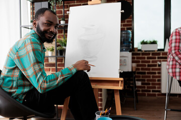 Art activities and wellbeing. Portrait of happy smiling African American guy during drawing lesson, young black man sitting at easel looking at camera, feeling inspired during sketching class