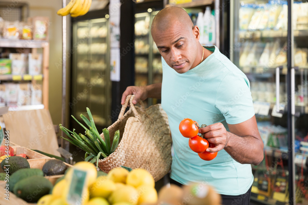 Wall mural man shopper chooses ripe tomatoes at grocery supermarket